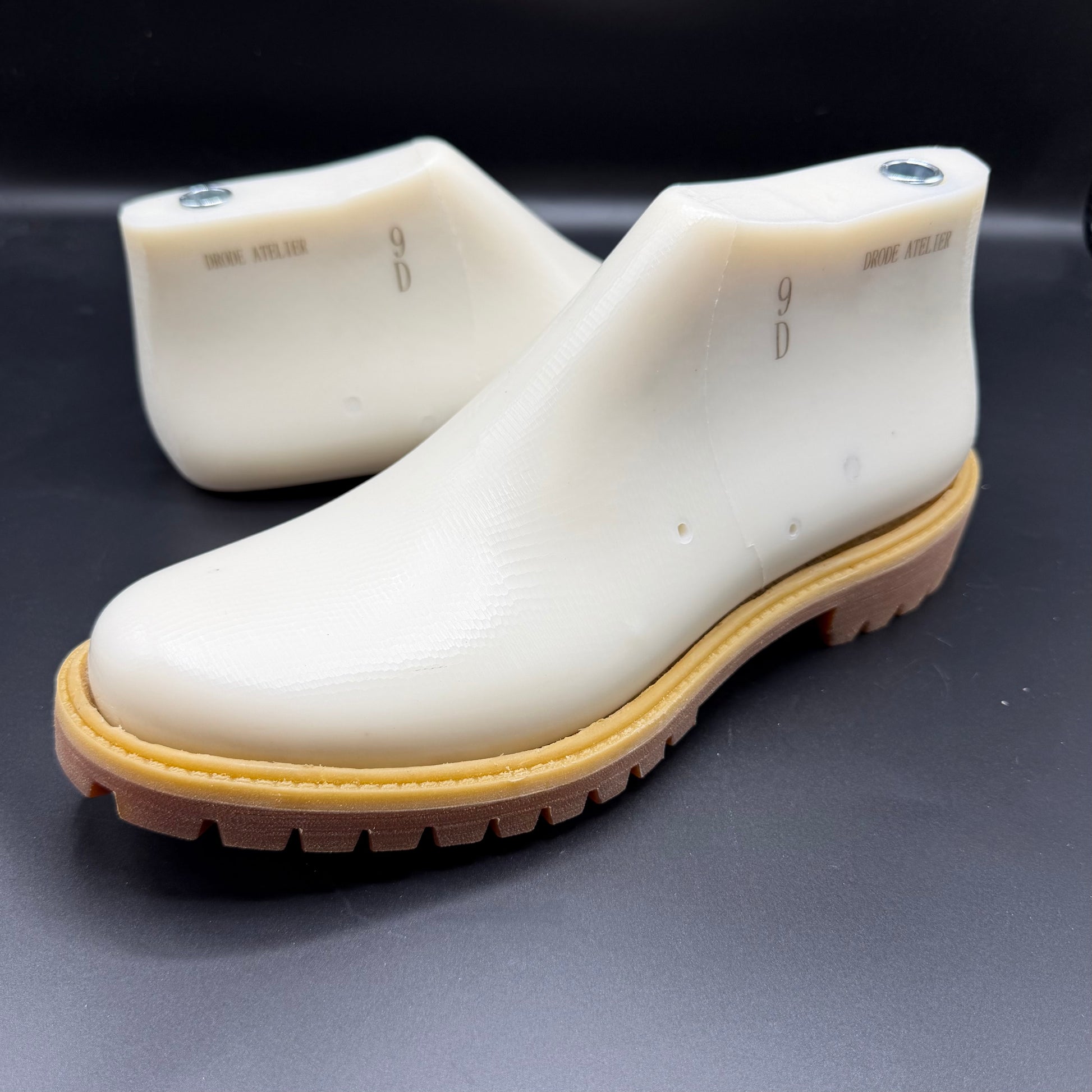 A pair of timb boot style shoe lasts with a tan and brown sole, displayed on a dark gray surface.